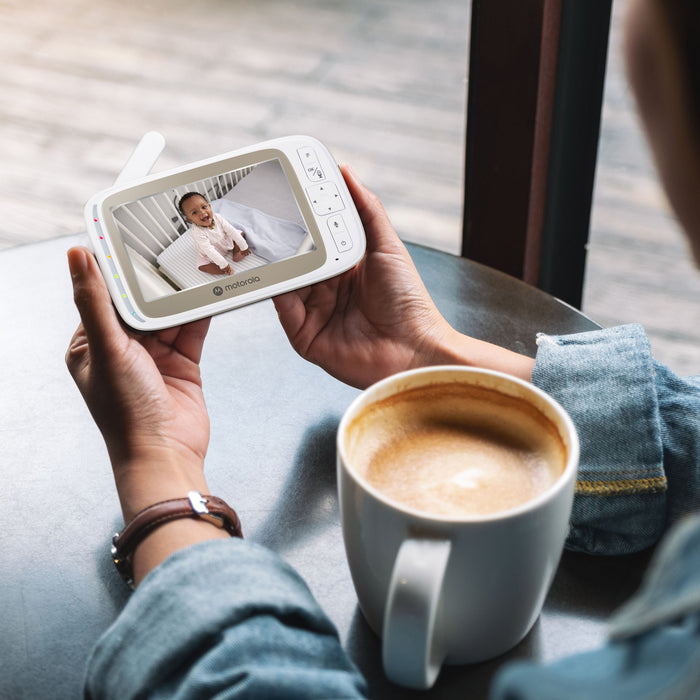 A person sitting at a table holds a handheld baby monitor showing a baby on the screen, with a cup of coffee in the foreground and a real indoor environment in the background.